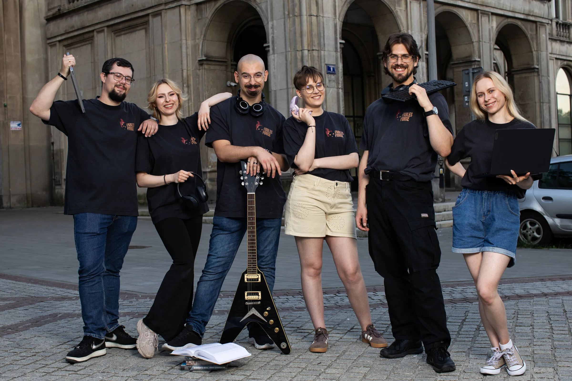 Zespół Fables Forge w studyjnych koszulkach, z gitarą, słuchawkami, klawiaturą i laptopem, przed budynkiem z arkadami